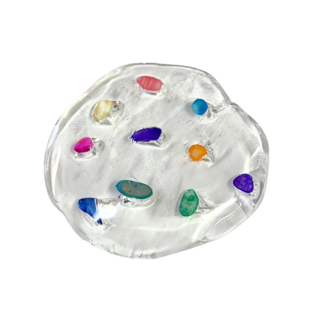 A circular blob of clear, translucent sensory putty containing various small, colorful tumbled stones and crystals in shades of pink, blue, green, and purple. The putty is shown against a plain white background, highlighting its use for fine motor skill development and tactile "pick-em" play.