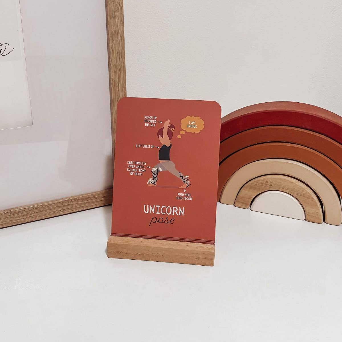 Red yoga card with text and illustration of unicorn pose on a wooden stand, next to wooden rainbow toys.