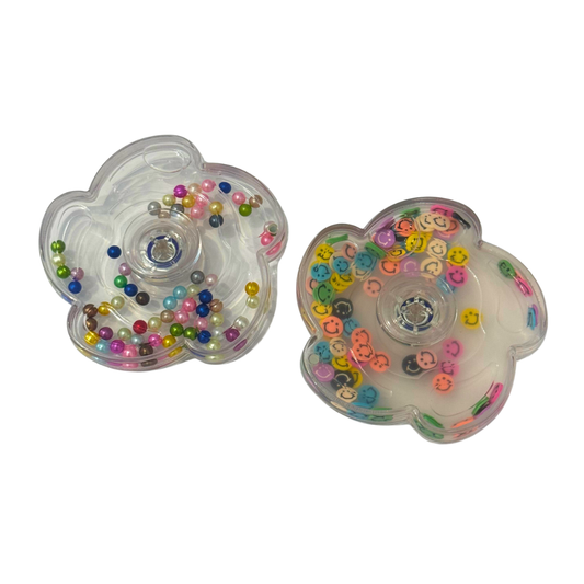 Two flower-shaped, clear plastic fidget spinners are shown side-by-side. The left spinner contains vibrant, metallic multi-colored beads, while the right spinner is filled with colorful smiley-face discs.