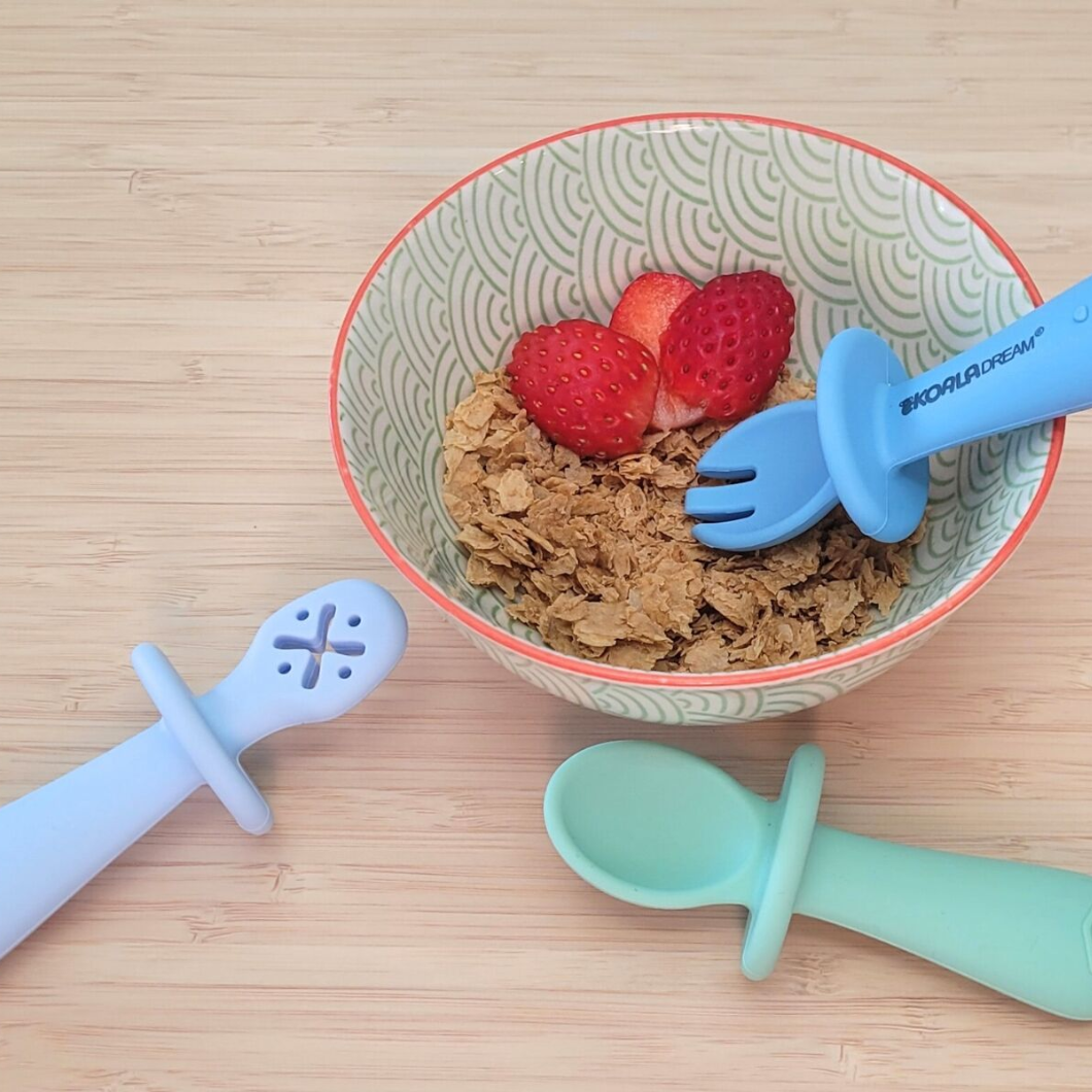 Bowl of cereal with strawberries and baby utensils on a wooden surface
