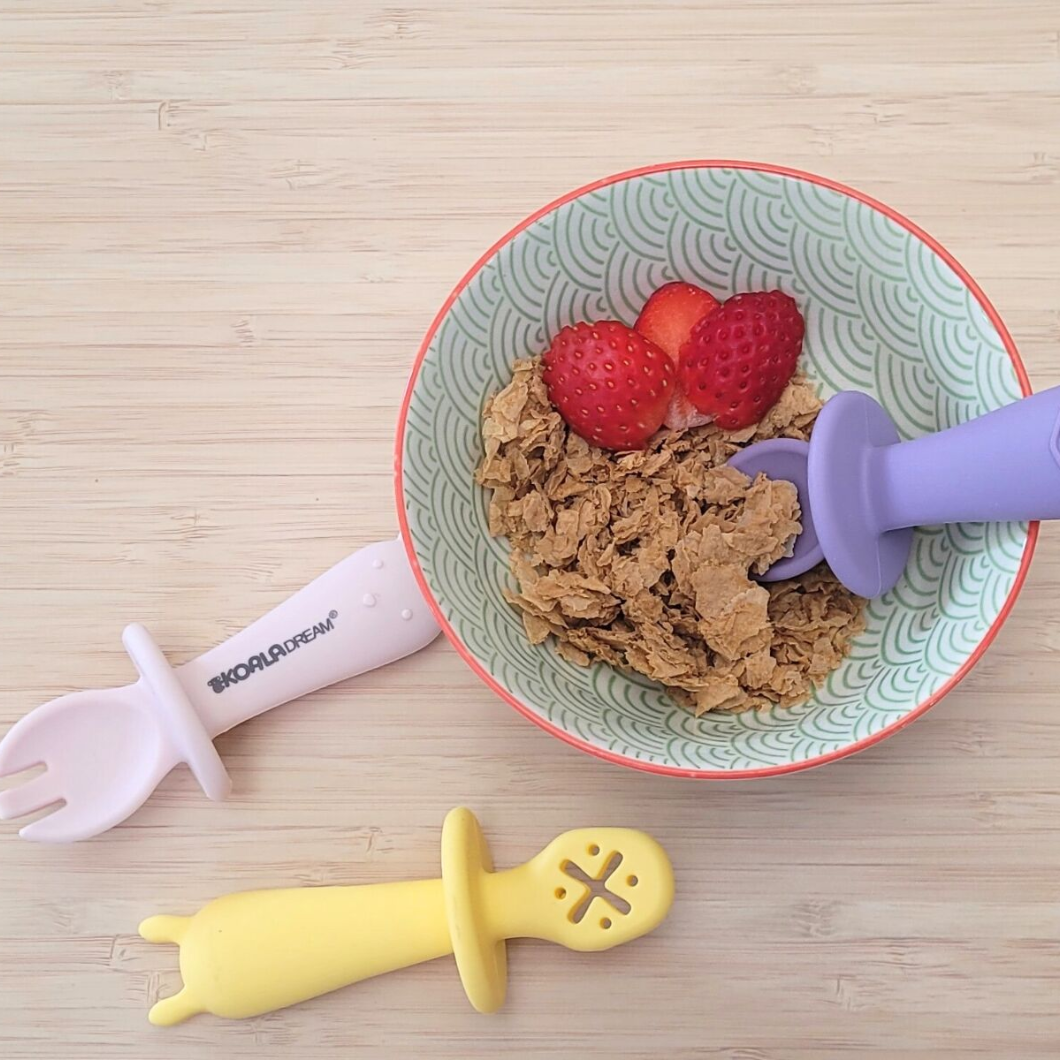 Bowl with cereal and strawberries, with baby utensils on a wooden surface.