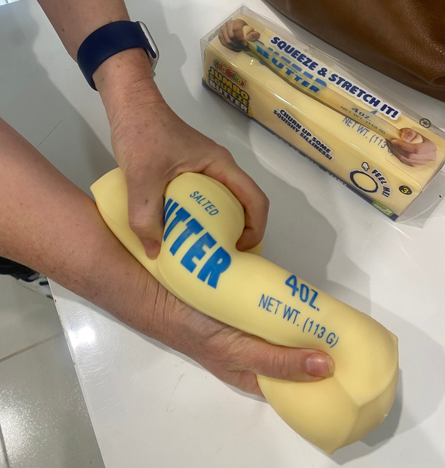 two hands squeezing the jumbo butter Squishy with a box of Jumbo Squeeze Butter by Smoosho in the background
