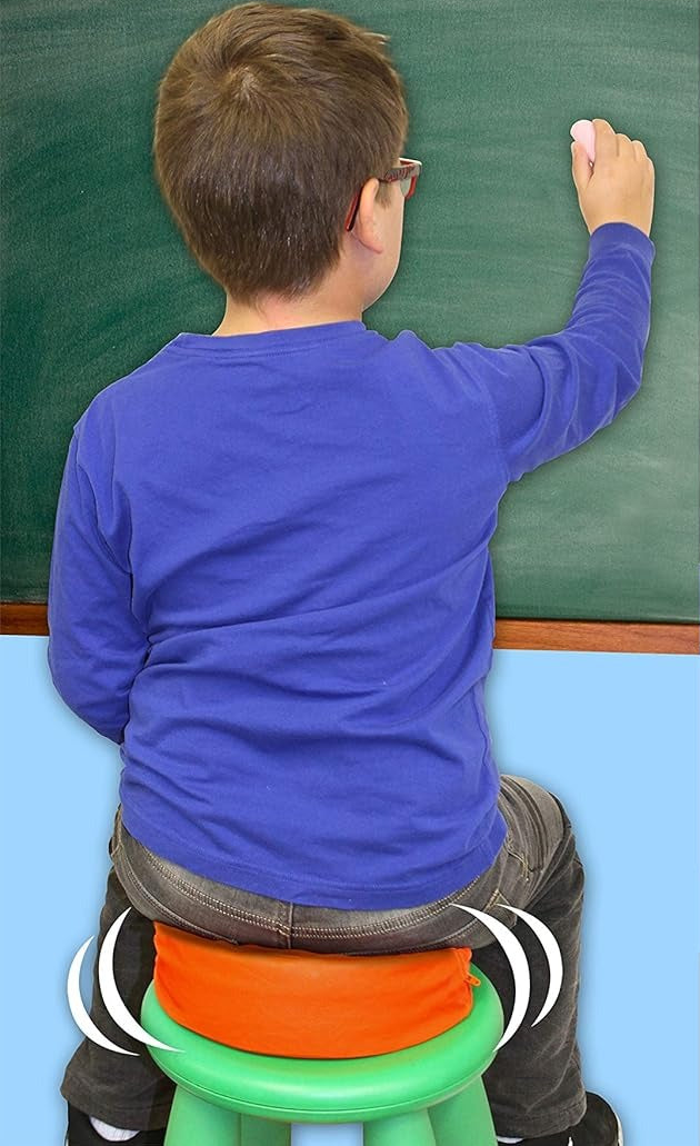 Child in a blue shirt sitting on an orange round vibrating cushion and writing on a green chalkboard.