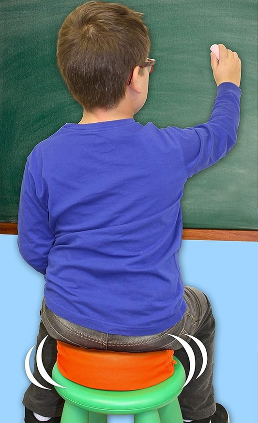 Child in a blue shirt sitting on an orange round vibrating cushion and writing on a green chalkboard.