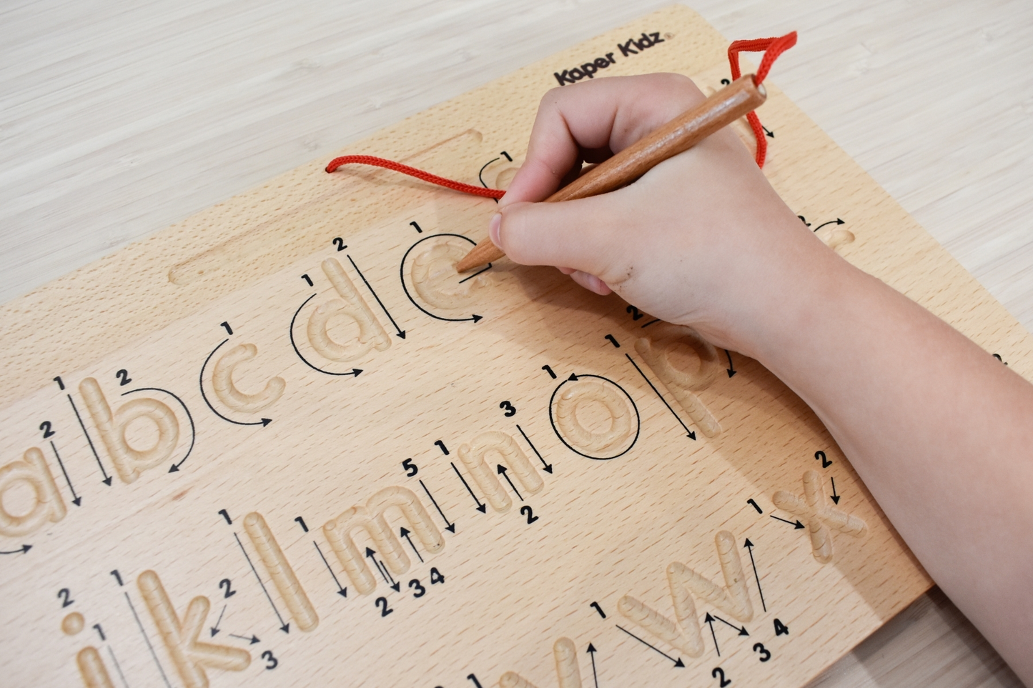 Child's hand using a wooden stylus to trace lowercase letters on a wooden tracing alphabet educational board.